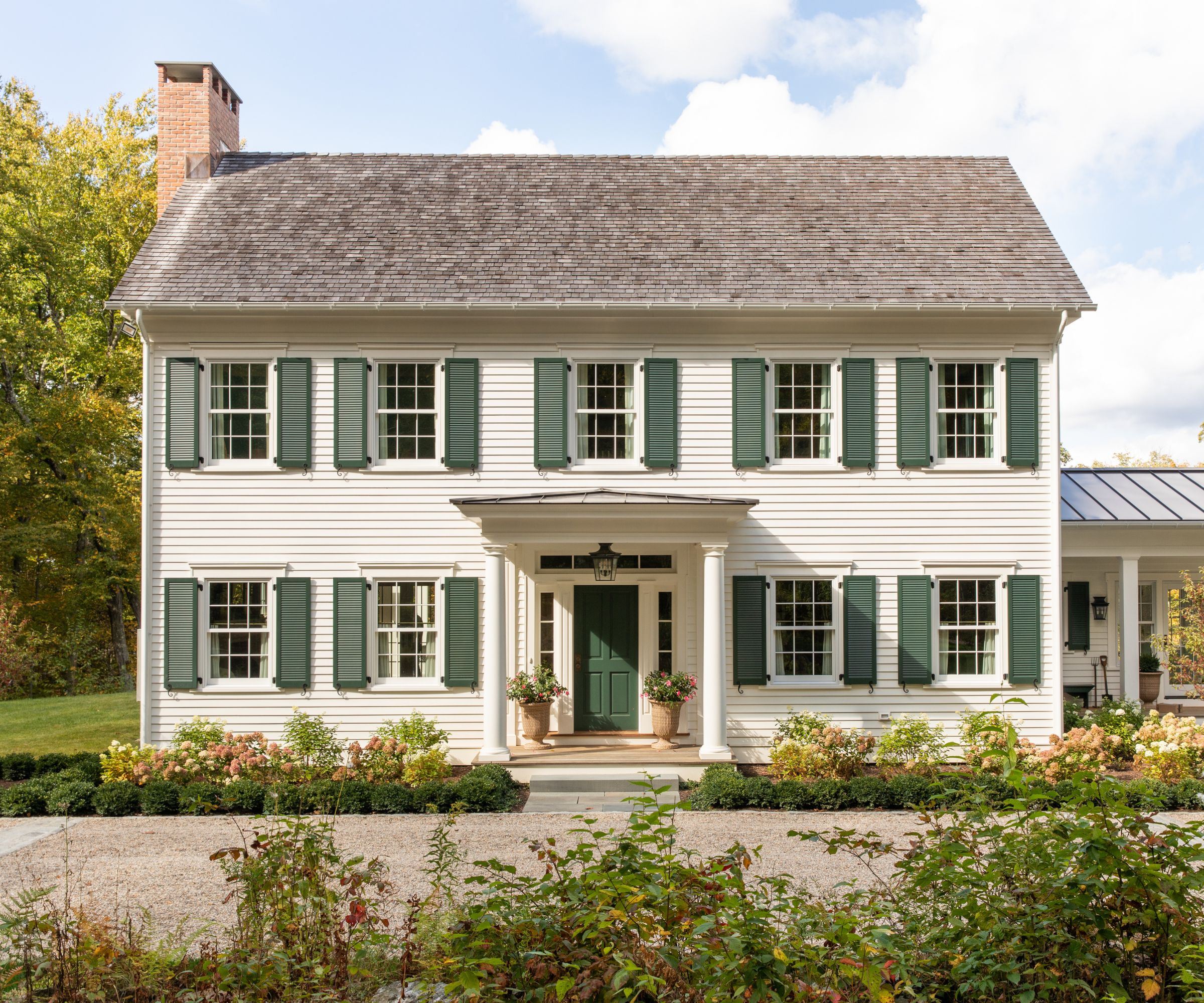 A large traditional house exterior with warm white siding, a dark green front door, and dark green shutters, with a porch and flower beds at the front of the home.