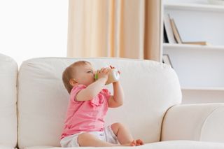 A baby sits on a couch, drinking from a bottle.