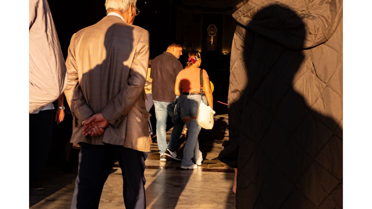 A man in a suit stands with hands behind his back, facing people walking ahead inside a sunlit building. Large shadows cover parts of the scene