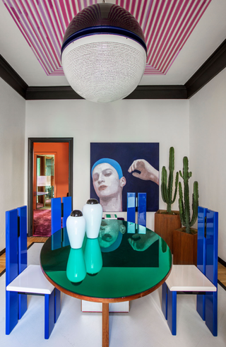 Image of a white and black dining room with a space-age-inspired design. There are four indigo dining chairs around a reflective cyan, oval dining table. The ceiling is painted in a pink-striped design and there is a black-and-white globe pendant light.