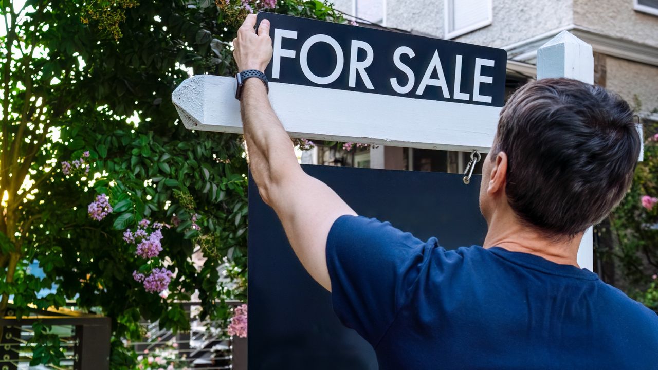 Close-up rear view of man adjusting &quot;For Sale&quot; sign in the front yard of a house