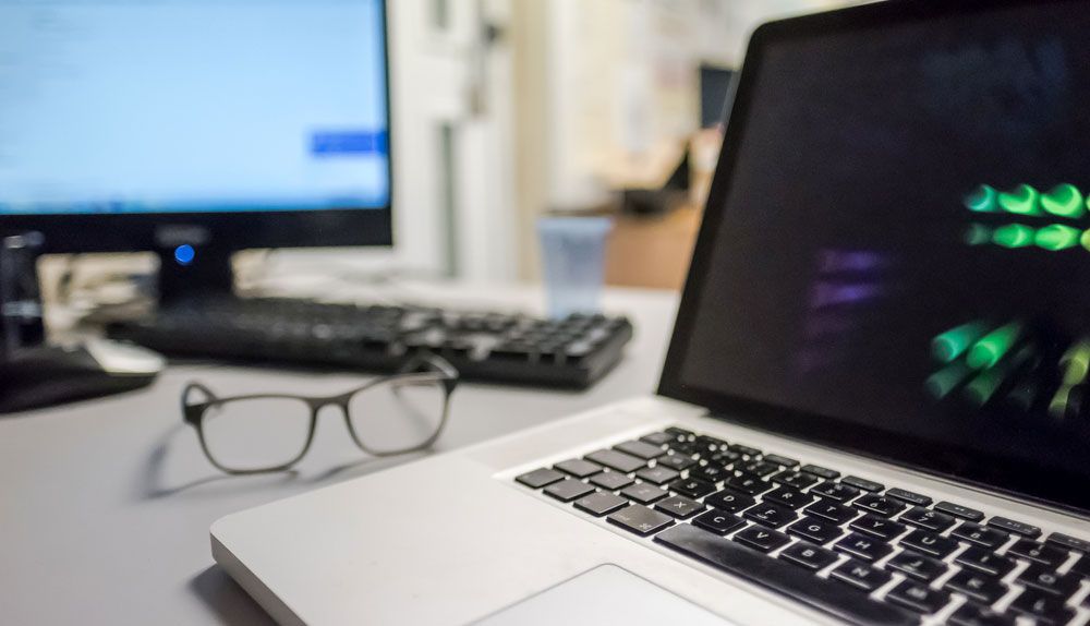 MacBook on desk with glasses and monitor displaying Windows in background.