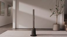 Shark TurboBlade fan in vertical position in neutral room atop white rug, in front of white-gray wall, large potted indoor tree, and hallway with two framed pictures.