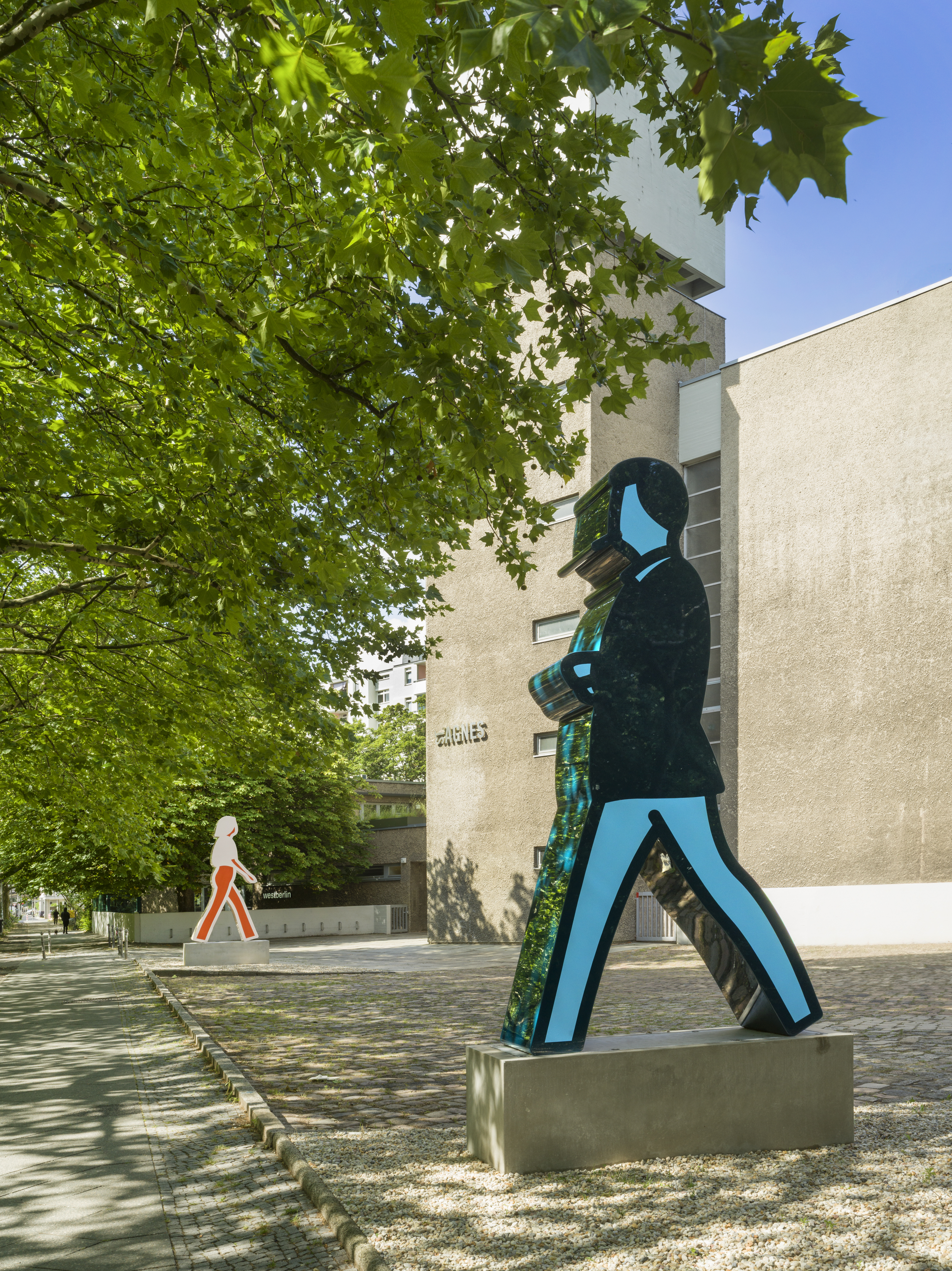 Two large-scale installations mimicking the semaphor-style silhouette of people walking, installed on a lush road with planes, natural light, and shadows reflecting on the pavement.