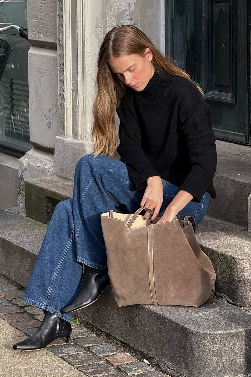 a woman wearing a black turtleneck, blue jeans, suede tote bag, and black kitten heel boots