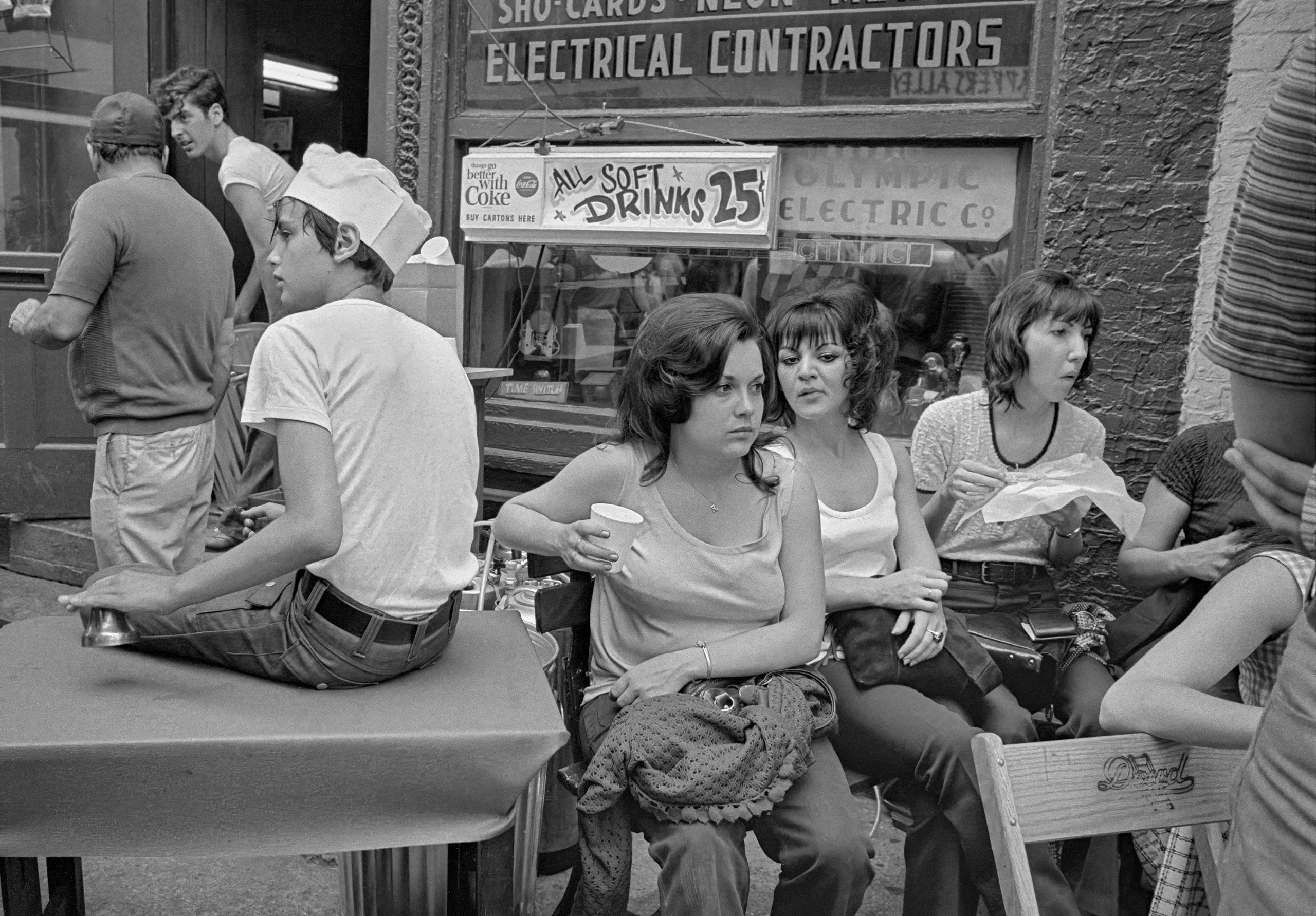Three young women sit on chairs outside a shop in Greektown beneath a hand-painted sign reading "All soft drinks 25&amp;cent;", with a boy in a paper hat perched on a table beside them.