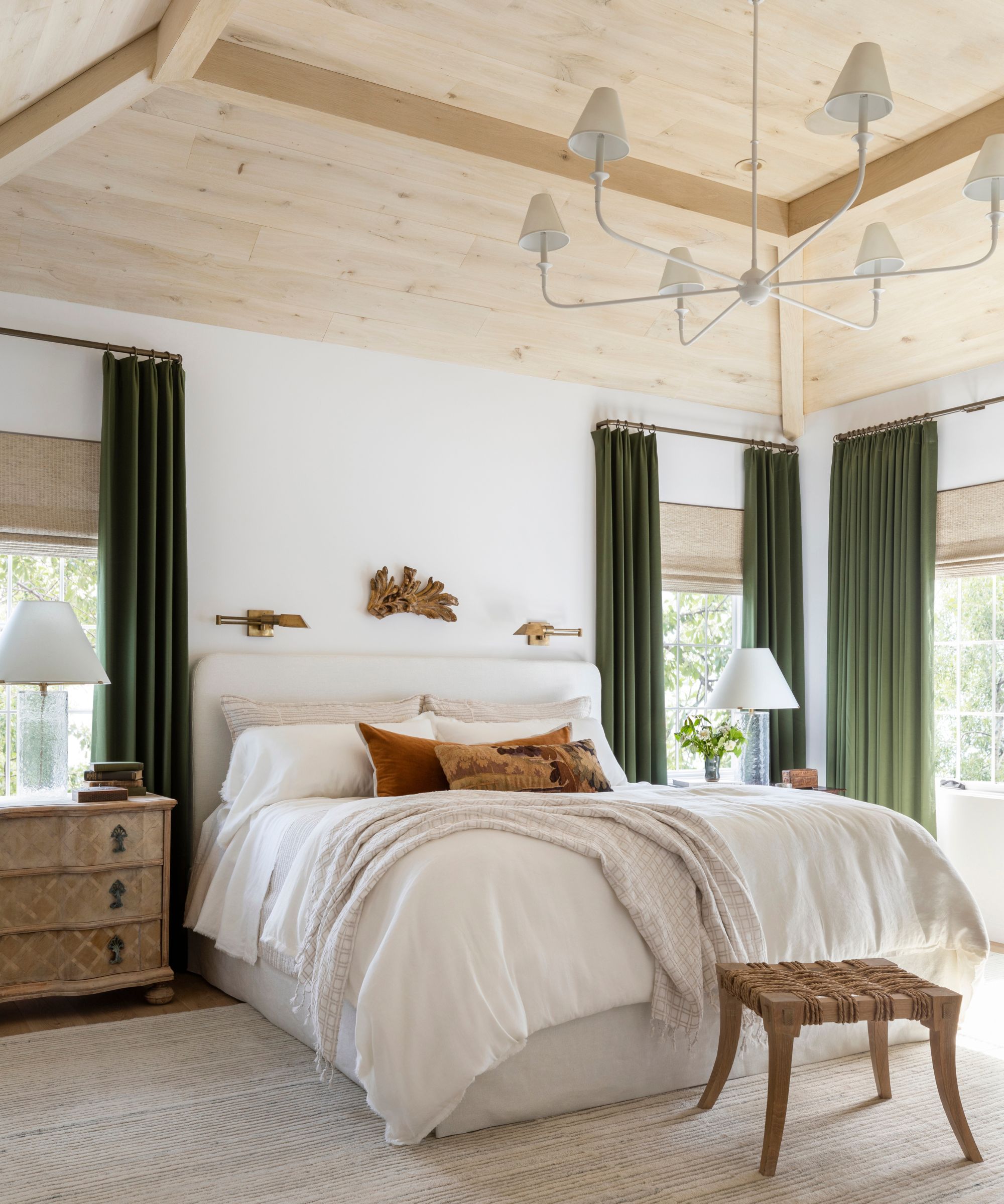 A modern light and airy bedroom with a pale wood-clad ceiling, white walls, a large beige rug, olive green curtains, and a large bed with layered neutral bedding.