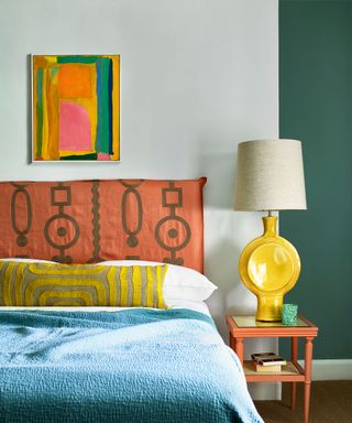 Colorful bedroom with white and green painted walls, colorful abstract artwork above bed, patterned, upholstered orange headboard, bed with white linen, yellow patterned cushion, blue throw, sculptural yellow table lamp with cream shade on orange bedside table
