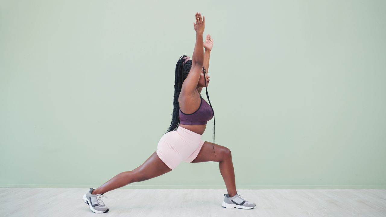 A woman in a sports bra, shorts and sneakers practices a yoga crescent lunge. Her left leg is bent at a right angle in front of her, while her right leg is extended behind. Her torso is upright and her hands reach towards the ceiling. 