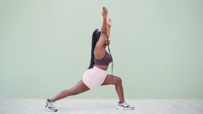 A woman in a sports bra, shorts and sneakers practices a yoga crescent lunge. Her left leg is bent at a right angle in front of her, while her right leg is extended behind. Her torso is upright and her hands reach towards the ceiling. 