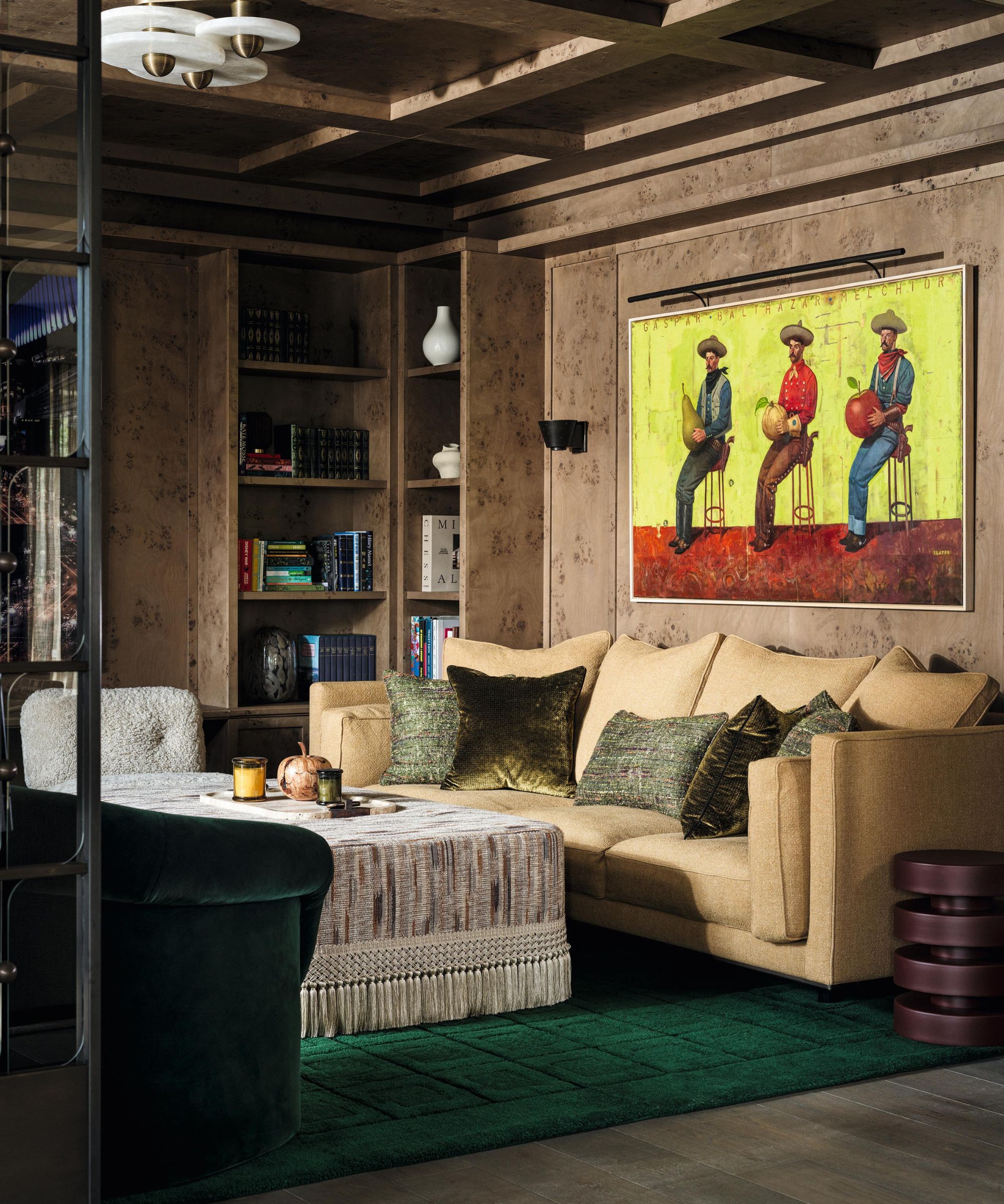 a burl wood clad sitting room with paneled ceiling, bespoke bookcases, a large green rug, caramel couch and statement cowboy painting