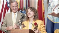 a woman in a yellow blazer and white blouse with brown wavy hair holds a red rose as she speaks into a microphone at a podium while a man in a tan jacket, white shirt and yellow tie with light brown hair looks on.