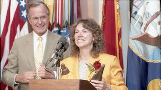 a woman in a yellow blazer and white blouse with brown wavy hair holds a red rose as she speaks into a microphone at a podium while a man in a tan jacket, white shirt and yellow tie with light brown hair looks on.