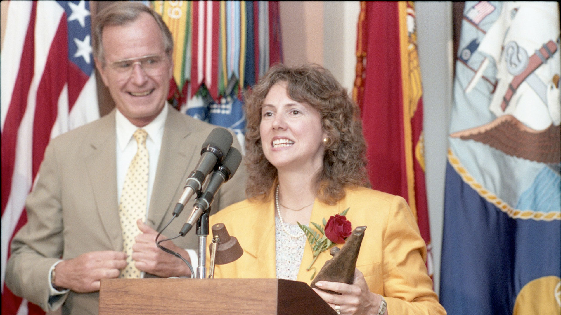 a woman in a yellow blazer and white blouse with brown wavy hair holds a red rose as she speaks into a microphone at a podium while a man in a tan jacket, white shirt and yellow tie with light brown hair looks on.