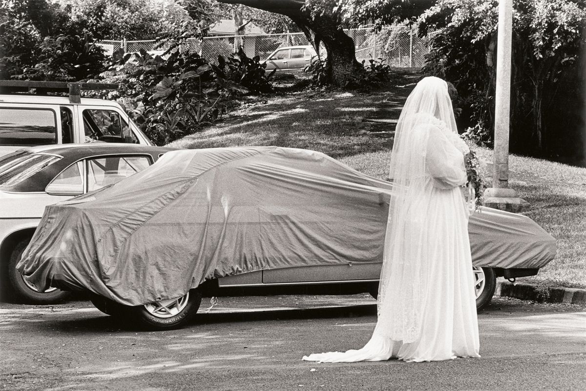 A black and white image showing bride in a white gown stands beside a car covered with a cloth