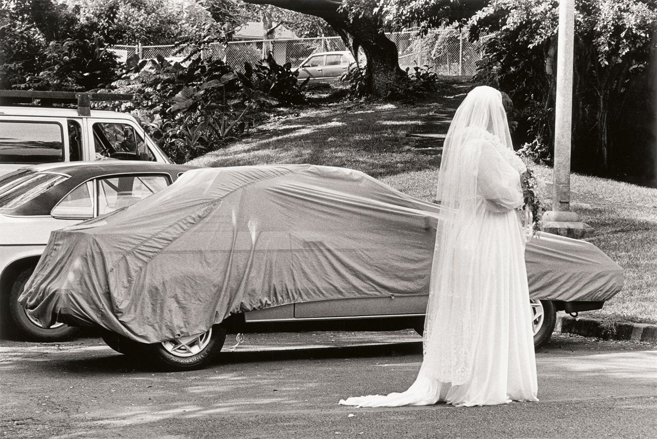 A black and white image showing bride in a white gown stands beside a car covered with a cloth