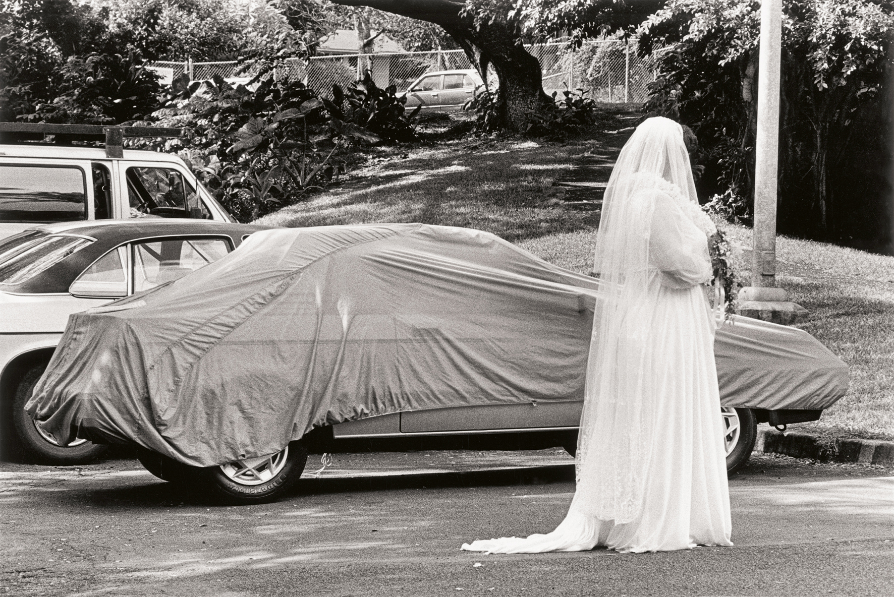 A black and white image showing bride in a white gown stands beside a car covered with a cloth