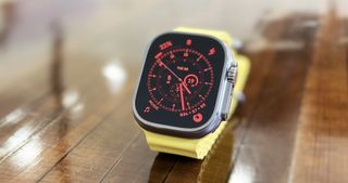 En Apple Watch Ultra med ett ljusgult armband står uppställd på ett träbord.