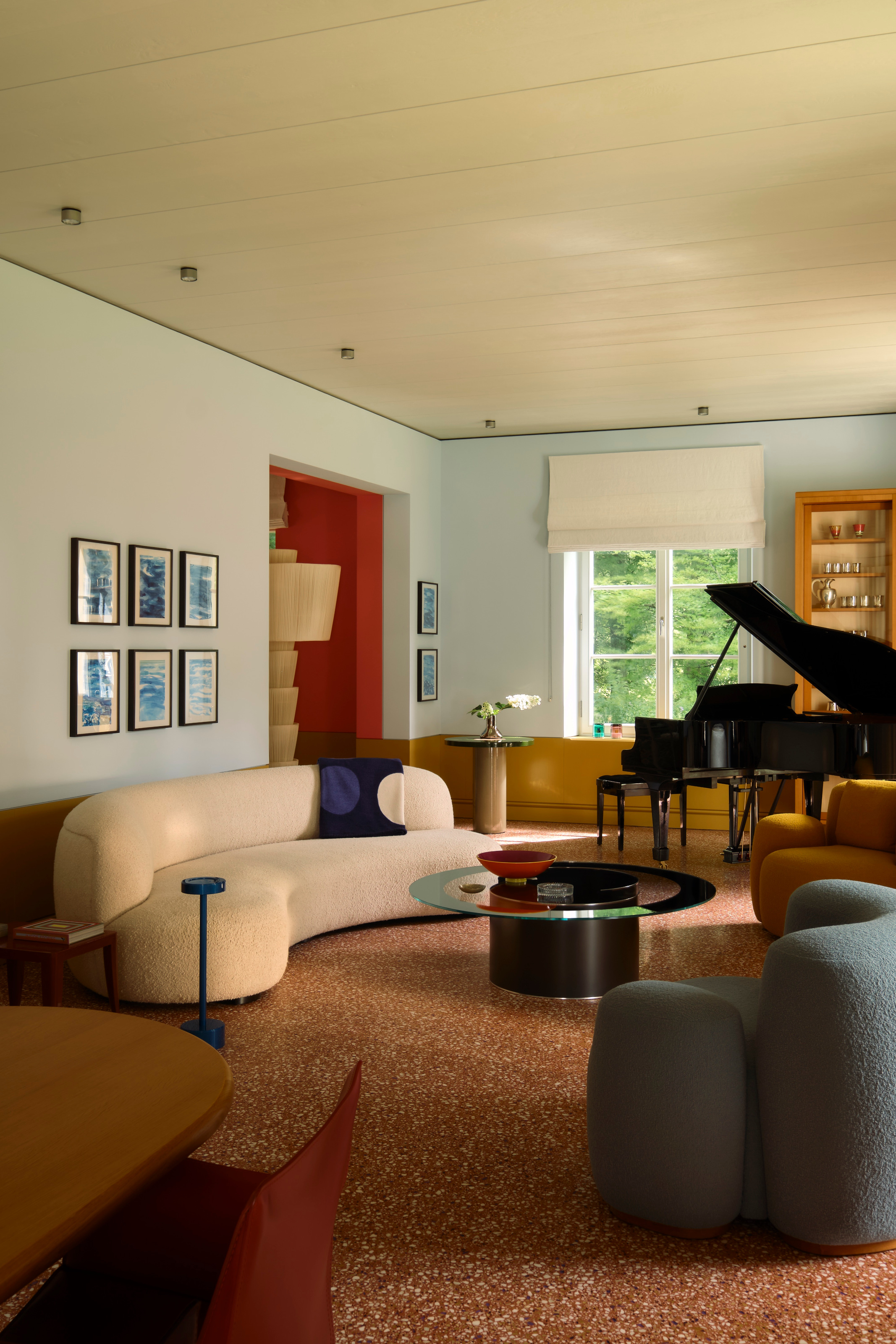 Living room with pale blue walls, red terrazzo floor, curved cream sofa and blue and mustard armchairs, glass round coffee table, black gran piano and dark blue cushion