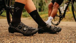 A close up of a riders black shoes. They are astride a bike on a muddy trail.