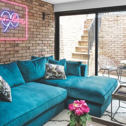 Basement living room with exposed brick wall, teal velvet sofa, neon light and large windows leading to steps and small courtyard area