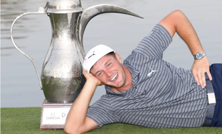 Bryson DeChambeau lies on his side in front of the Dubai Desert Classic trophy in 2019