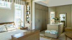 Neutral large bedroom on left with walk in closet area on the right. The closet has built in floor to ceiling closets visible, as well as an island with drawer storage. The natural decor is calming and neutral, with stripes and blue accents