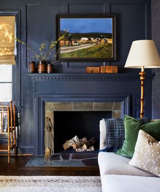 inky blue living room with paneling and fireplace painted the same color, cream couch, bamboo floor lamp, artwork, hardwood floor, blind