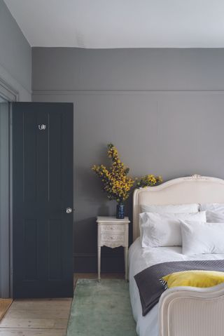 grey bedroom with pale green rug, white French style bed, matching side table, yellow flowers, yellow cushion, dark grey painted door