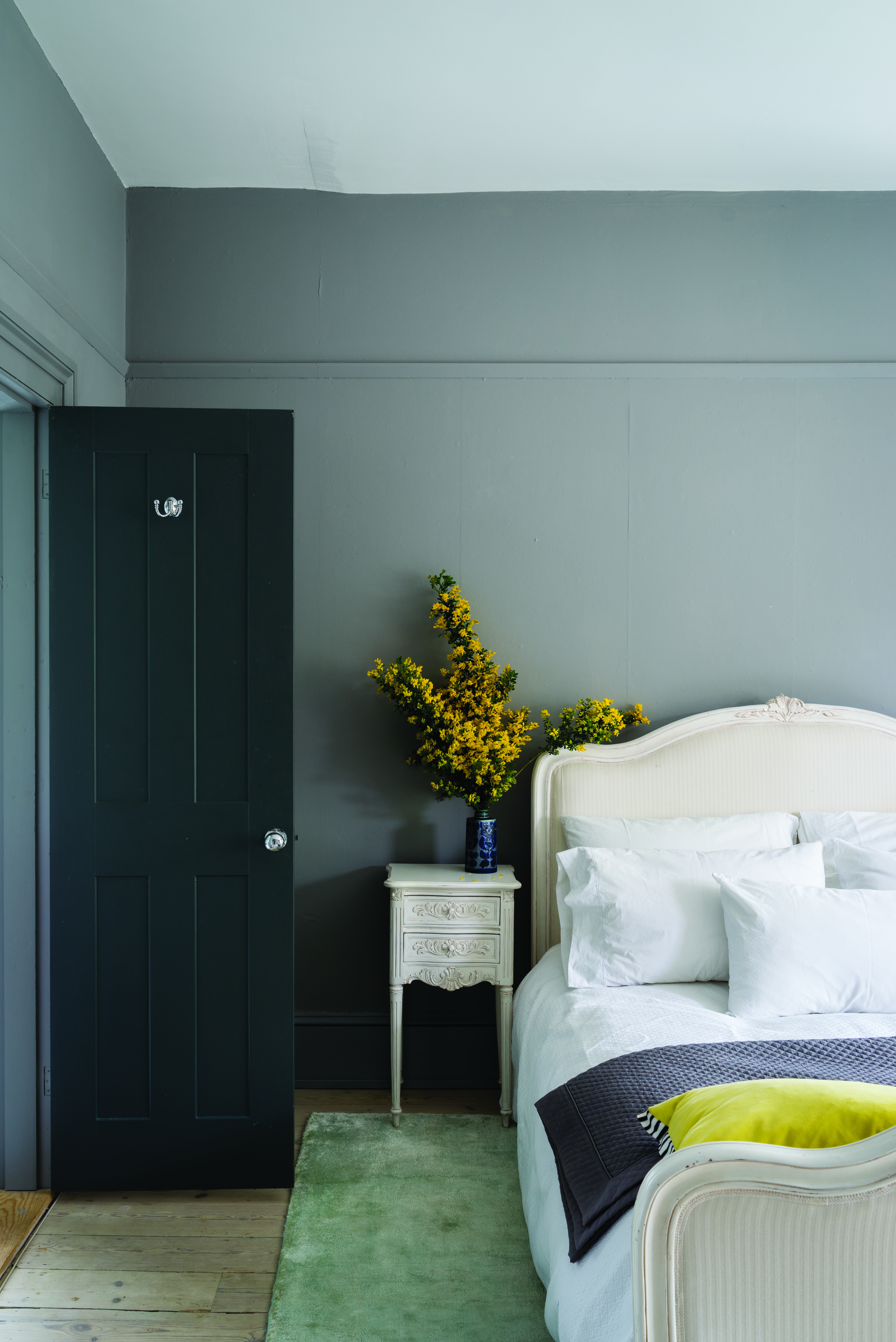 grey bedroom with pale green rug, white French style bed, matching side table, yellow flowers, yellow cushion, dark grey painted door