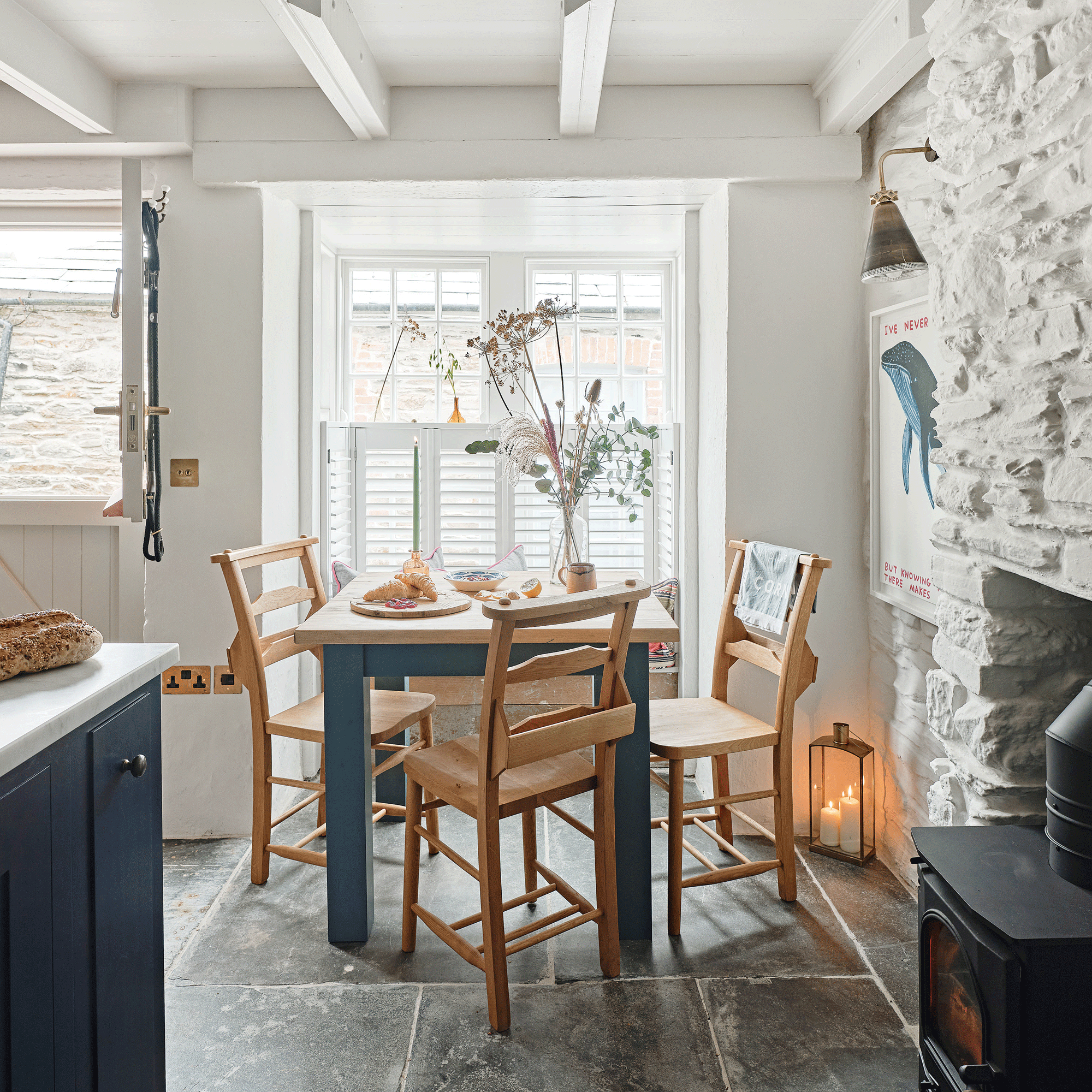 a dining area beside a kitchen in an open plan area beside a wood burning stove, a window with half height shutters