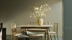 a wooden dining table and chairs with vase of dainty flowers against a beige/gray wall