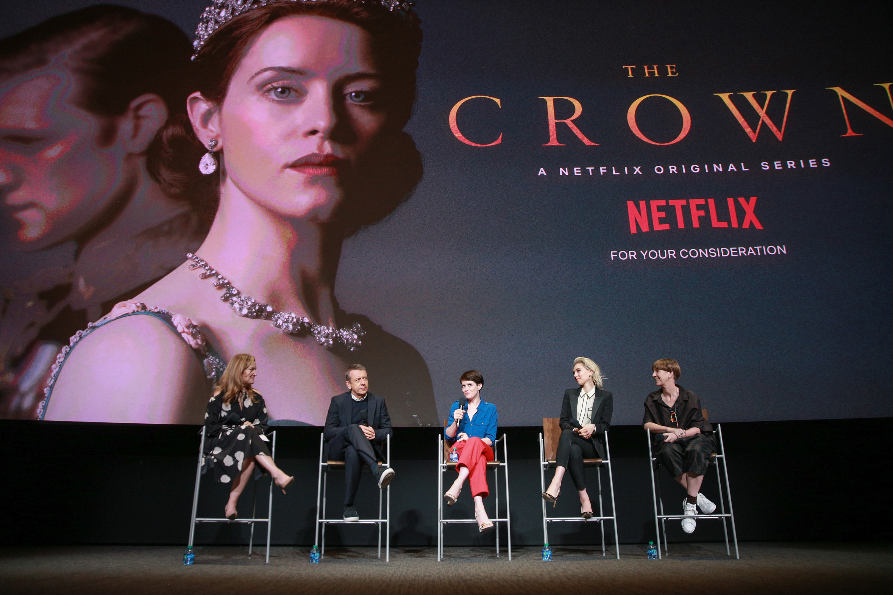 Cast members of The Crown sitting on stage in front of a show poster
