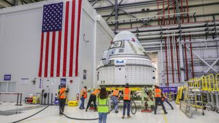 technicians surround a white space capsule in a large room, with an american flag on one wall