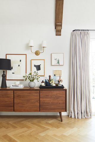 A wooden credenza in a bright room, topped with a bar tray, flowers and black lamp