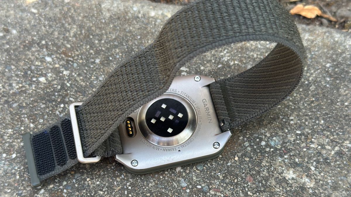 The Garmin Venu X1 sitting upside-down on asphault, with the words "Titanium &bull; Venu X1 &bull; Sapphire Crystal &bull; Taiwan" visible around the health sensors, as well as the four-pin charging port and the nylon strap.
