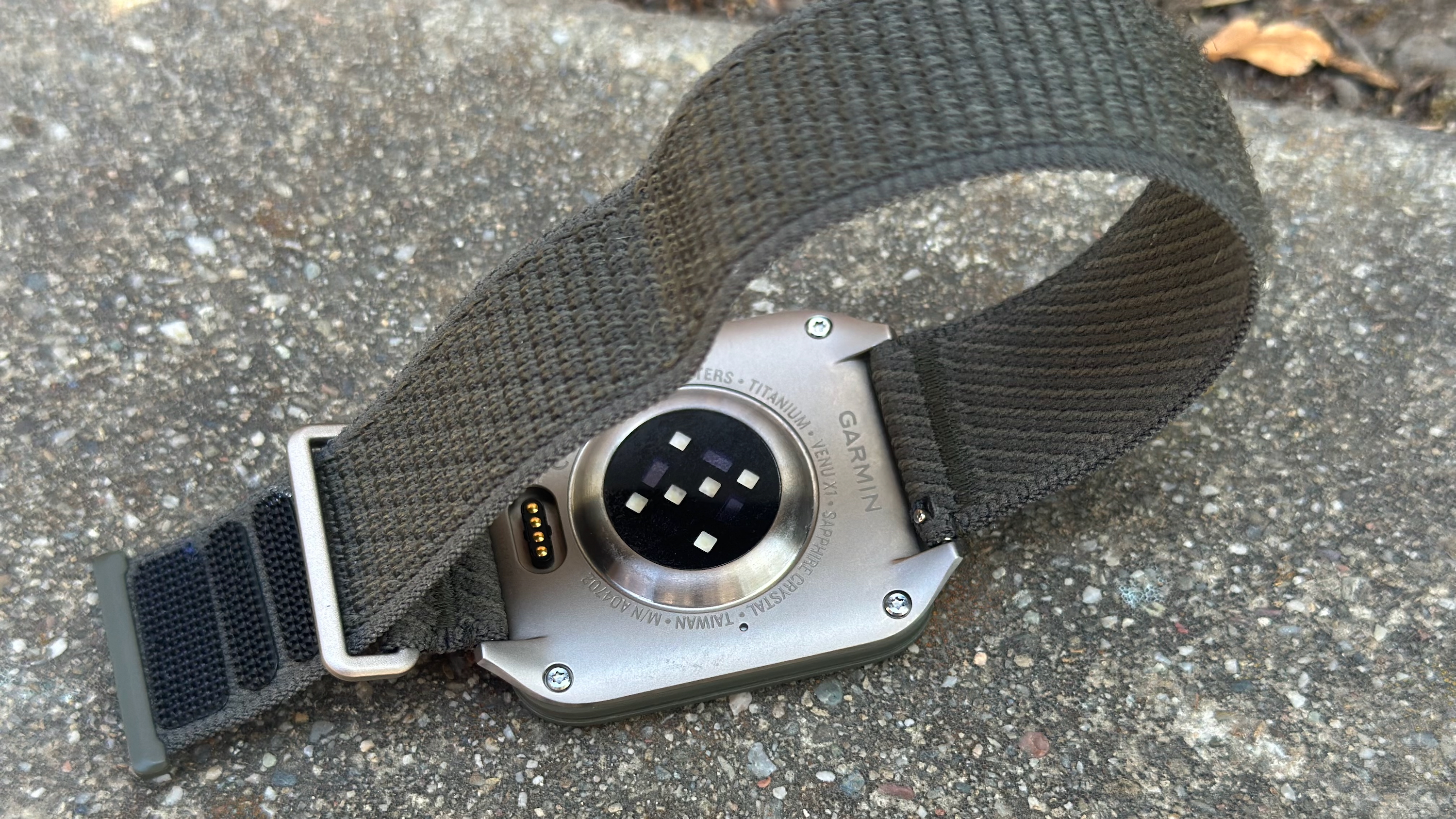The Garmin Venu X1 sitting upside-down on asphault, with the words "Titanium • Venu X1 • Sapphire Crystal • Taiwan" visible around the health sensors, as well as the four-pin charging port and the nylon strap.