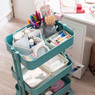 a small green movable trolley in front of a white desk, with three shelves containing an assortment of craft items
