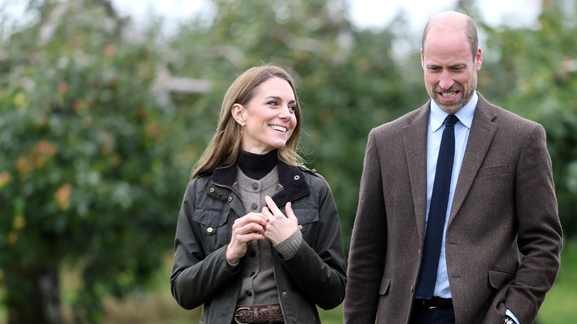 Kate Middleton wearing a green coat and brown cardigan walking next to Prince William in an orchard