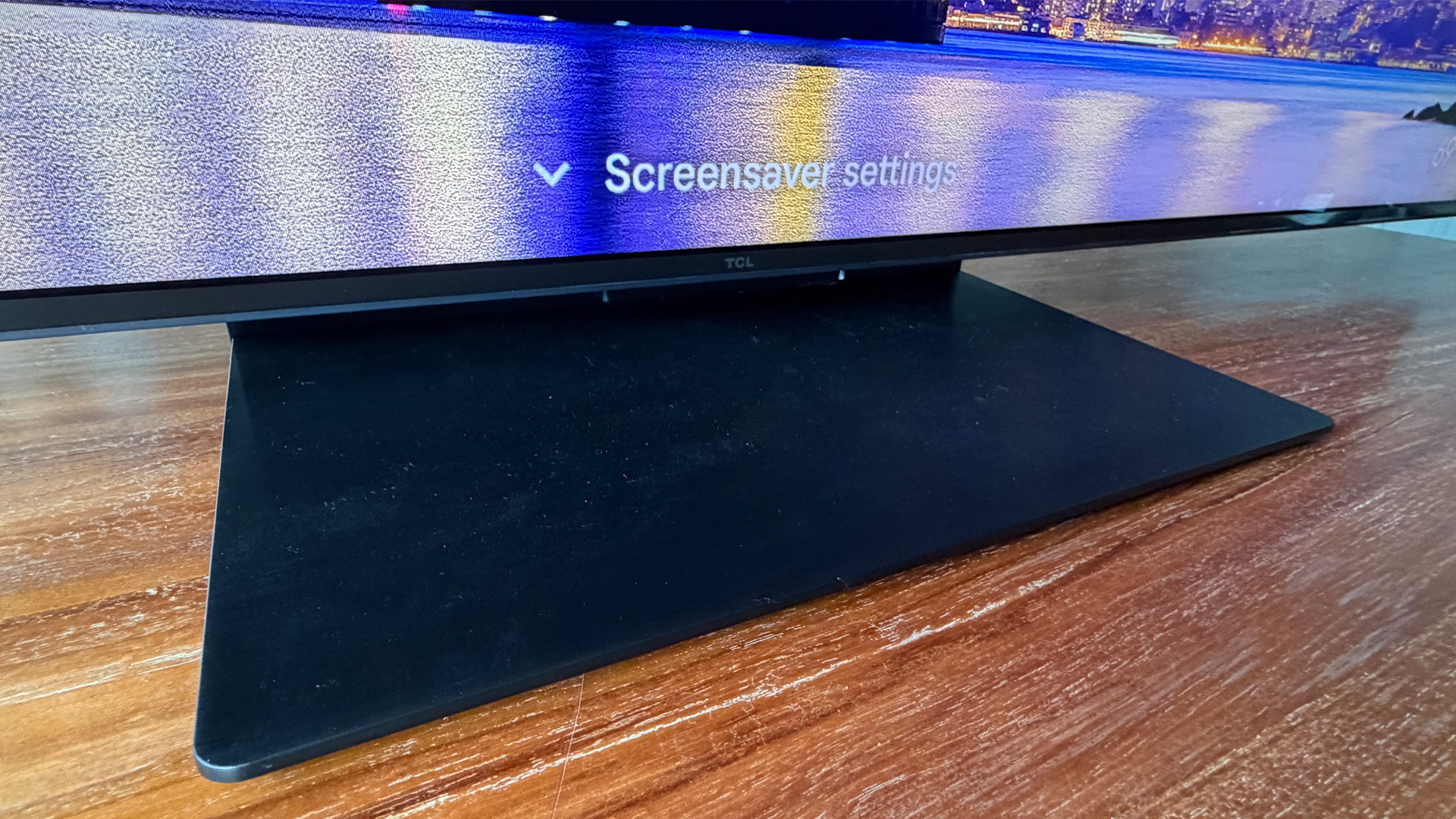 TCL C7L Mini LED TV on wooden table detail of pedestal stand