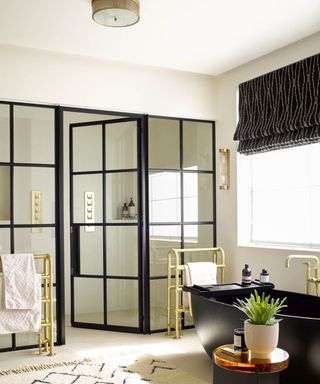 A white bathroom with black framed glass doors, a black matte bathtub and Venetian style black blinds, and a low-profile round gold ribbed ceiling light