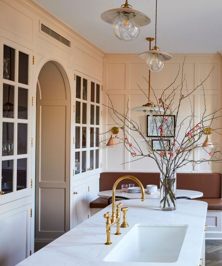 A pale pink kitchen with a curved island, brown banquette, and glass cabinets connected to the dining room