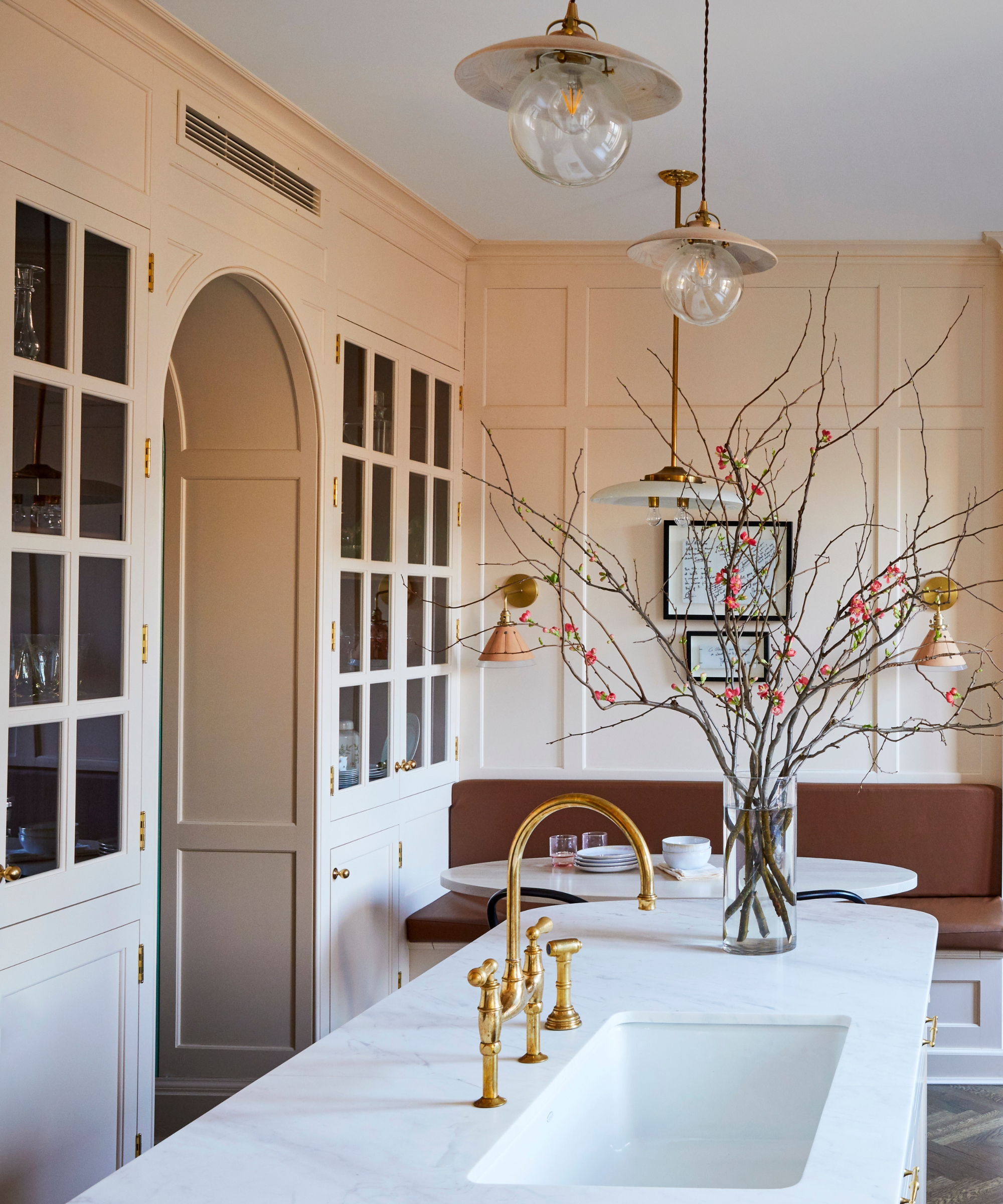 A pale pink kitchen with a curved island, brown banquette, and glass cabinets connected to the dining room