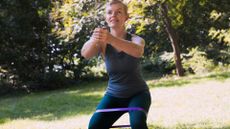 woman in dark coloured vest and leggings performing a half squat with a resistance band around her legs above her knees, outside in a park setting. 