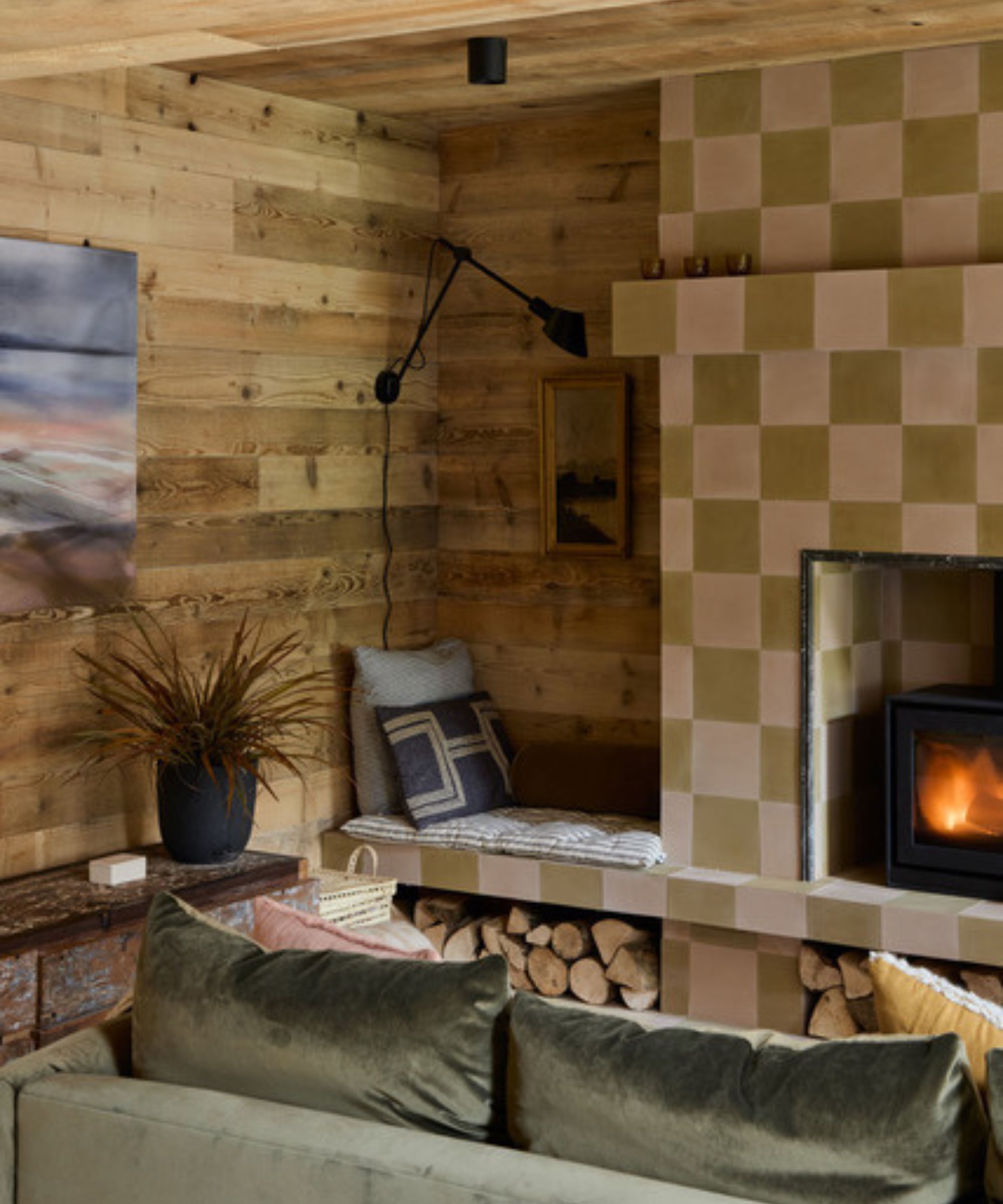 A rustic living area with timber-clad walls, a tiled fireplace in soft green and beige, and a wood-burning stove. A built-in bench with cushions sits beside stacked logs, creating a cosy, natural atmosphere with layered textures.