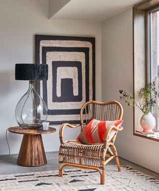 Sitting corner by large window, with large, striking, mono wall art, shapely cane chair with diagonal striped cushion, and sculptural round wood side table with large glass, shapely table lamp and black shade.