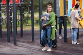 Laura (Jo Joyner) stands in a playground with baby Elliot strapped to her chest, looking around in panic