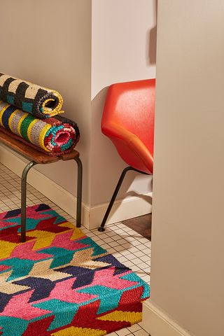An orange chair, two rolled up multicoloured Colville Jute floor mats on a small wooden bench and one floor mat laid out on the floor displaying its striped and arrowhead design