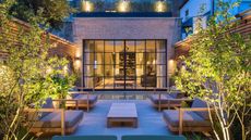 An urban modern garden, with a view of the home through the crittal style doors, with two large arm chairs either side of a garden coffee table with lit trees and fence panels all around