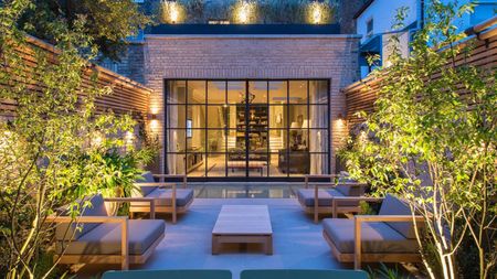 An urban modern garden, with a view of the home through the crittal style doors, with two large arm chairs either side of a garden coffee table with lit trees and fence panels all around